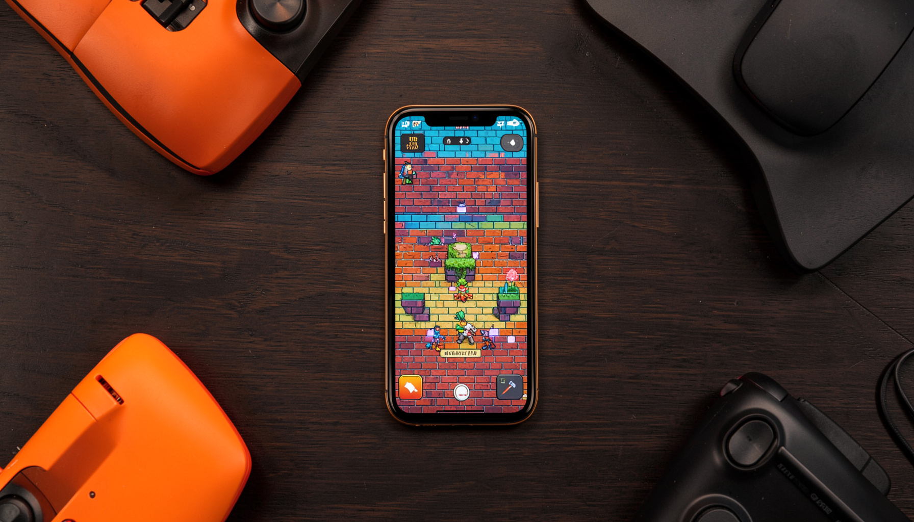 Overhead flat-lay of a smartphone displaying a colorful brick-wall descent game, surrounded by gaming accessories on a dark wooden desk, warm amber and charcoal tones.